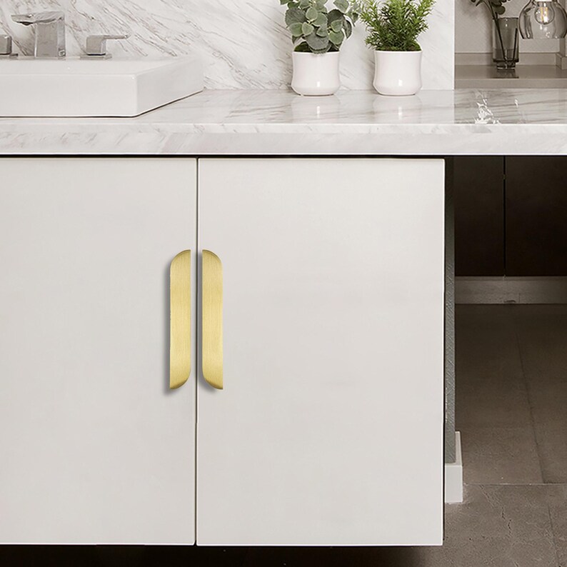 May include: A white cabinet featuring two vertical, gold-coloured handles. The handles have a curved design. The cabinet is set in a bathroom, with a white countertop and small potted plants visible in the background.