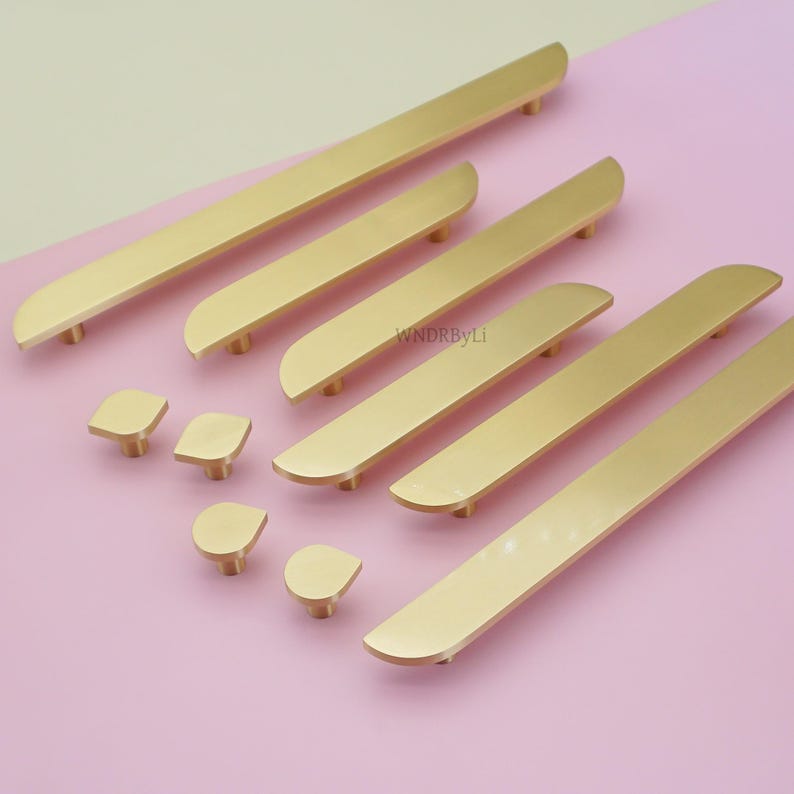 May include: A selection of gold-coloured cabinet hardware, including long bar pulls and square and teardrop-shaped knobs. The hardware is arranged on a pink surface, with the text "WNDRByLi" visible.
