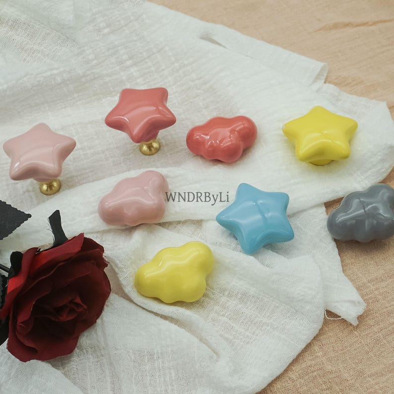 May include: A selection of ceramic cabinet knobs in star and cloud designs. The knobs are in various colours, including pink, yellow, blue, and grey, with gold-tone fixings. The text "WNDRByLi" is visible on the fabric.