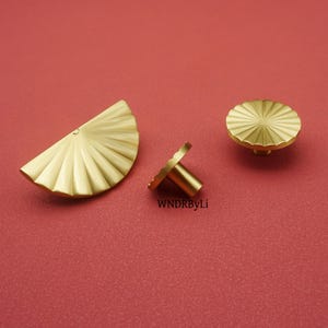 May include: Three gold-colored cabinet hardware pieces on a red background. The pieces include a half-circle pull, a knob, and a small handle. The hardware has a ridged, fan-like design. The text "WNDRByLi" is visible.