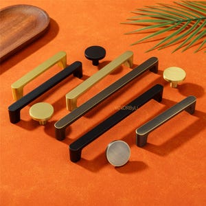 May include: A selection of cabinet handles and knobs in gold, black, and bronze finishes. The hardware is displayed on an orange surface, showing different shapes and styles. The image includes both bar pulls and circular knobs.