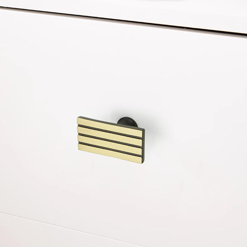 Puede incluir: Primer plano de un armario blanco con un tirador rectangular. El tirador es negro con tres rayas horizontales doradas. El tirador est&aacute; fijado al armario con una peque&ntilde;a base redonda negra.