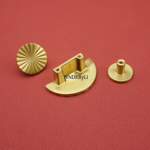 May include: Three brass cabinet hardware pieces on a red background. The set includes a round knob with a sunburst design, a half-moon shaped pull with two screw holes, and a small round knob with a cylindrical post. The text "WNDRByLi" is visible on the pull.