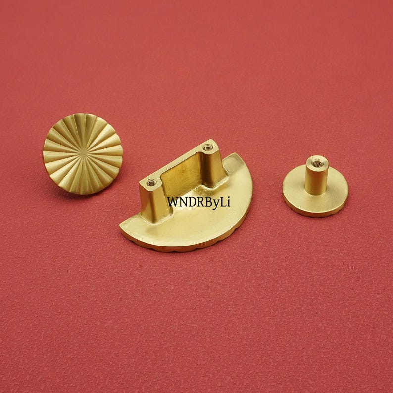 May include: Three brass cabinet hardware pieces on a red background. The set includes a round knob with a sunburst design, a half-moon shaped pull with two screw holes, and a small round knob with a cylindrical post. The text "WNDRByLi" is visible on the pull.