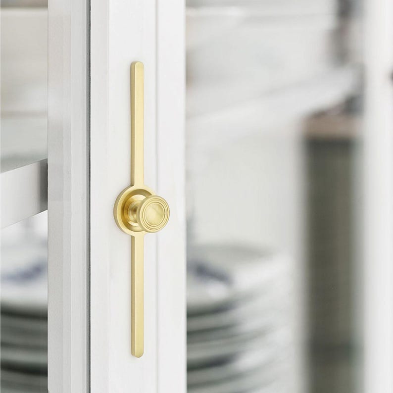 May include: A close-up of a gold-coloured cabinet latch. The latch is mounted on a white cabinet door. The latch has a long, slender handle and a circular knob with a ridged design.