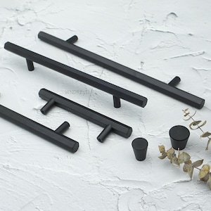 May include: A collection of matte black cabinet hardware, including bar pulls and knobs. The pulls have a hexagonal design and are available in various lengths. The knobs are cylindrical and conical. The hardware is displayed on a textured white surface.
