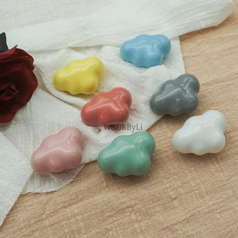 May include: Seven glossy ceramic cloud-shaped ornaments in pink, yellow, red, blue, grey, green, and white. The decorative pieces are arranged on a white cloth. The text "WNDRByLi" is visible.