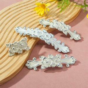 May include: A collection of white and silver floral cabinet pulls. The pulls feature a detailed floral design with a silver outline. The hardware is arranged on a wooden surface, showcasing the decorative elements.