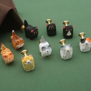May include: A selection of hand-carved wooden cat-shaped cabinet knobs. Each knob features a unique cat design with painted details in various colours, including orange, black, white, and yellow. The knobs have gold-coloured metal stems.