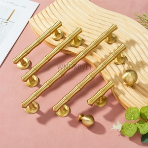 May include: A selection of gold-coloured bamboo-style cabinet hardware, including handles and knobs. The handles are long and cylindrical with a segmented design, while the knobs are spherical. The hardware is displayed on a wooden surface and a pink background.