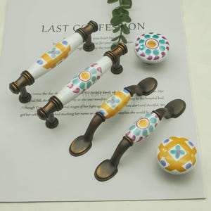 以下が含まれることがあります： 引き出しプルとノブを含む、装飾的なキャビネットハードウェアのコレクション。 プルは、カラフルな花の模様とブロンズトーンの金属ベースを備えた白いセラミックハンドルを備えています。 ノブは同様のパターンで丸いです。