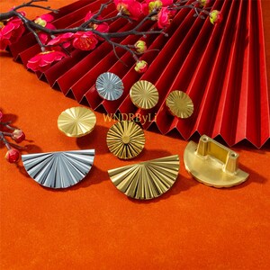 May include: An assortment of decorative hardware, including gold and silver cabinet knobs and drawer pulls, arranged on an orange surface with a red fan and floral accents. The hardware features a sunburst design.