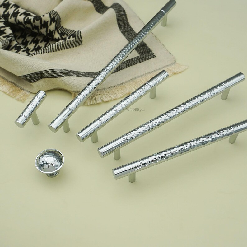May include: A collection of silver cabinet hardware, including several bar pulls, a small knob, and a T-shaped pull. The hardware features a hammered texture on the main body and smooth, cylindrical ends. The hardware is displayed on a light-colored surface.