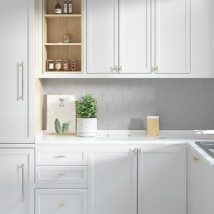 Op de afbeelding: Een lichte, moderne keuken met witte kasten en gouden hardware. Een kleine plant in een pot en een ingelijste print met de woorden "ME YOU" staan op het aanrecht. De achterwand heeft een geometrisch tegelontwerp.