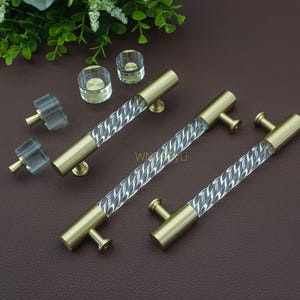 May include: Cabinet hardware set featuring brass-toned handles with clear, twisted acrylic centre sections. The set includes several knobs and handles, showcasing a modern design. The hardware is designed for furniture and home decor.