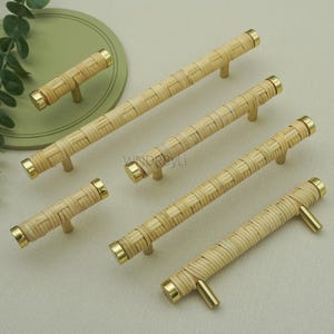 Könnte beinhalten: Eine Sammlung von gewebten Schrankgriffen aus Rattan mit messingfarbenen Endkappen und Griffen. Die Griffe sind unterschiedlich groß und auf einer neutralen Oberfläche angeordnet, wodurch ihre natürliche Textur und warmen Farbtöne zur Geltung kommen. Diese dekorativen Beschläge verleihen einen Hauch von rustikaler Eleganz.