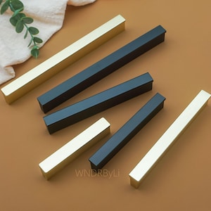 May include: A selection of rectangular cabinet pulls in gold and black. The pulls are arranged diagonally on a brown surface. The text "WNDRByLi" is visible in the lower right corner.