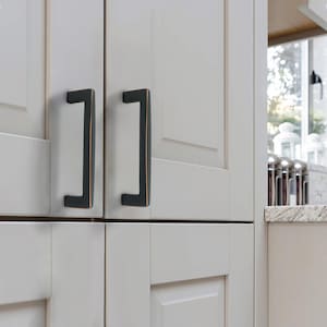 May include: Close-up of two dark bronze cabinet pulls on white cabinet doors. The pulls have a rectangular shape with a bronze accent on the edges. The cabinet doors are part of a kitchen cabinet set.