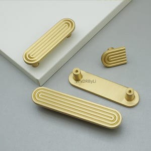 Op de afbeelding: Goudkleurige kastbeslagset. De set bevat verschillende rechthoekige handgrepen met een groefontwerp, een kleine knop en een rechthoekig stuk met twee schroefgaten. De hardware is gemaakt van metaal en heeft een matte afwerking.