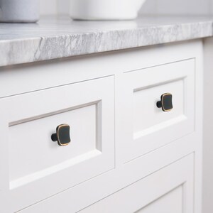 May include: Close-up of white cabinet doors with square black and gold cabinet knobs. The knobs have a dark, matte finish with a gold trim. The cabinet doors are set against a marble countertop.