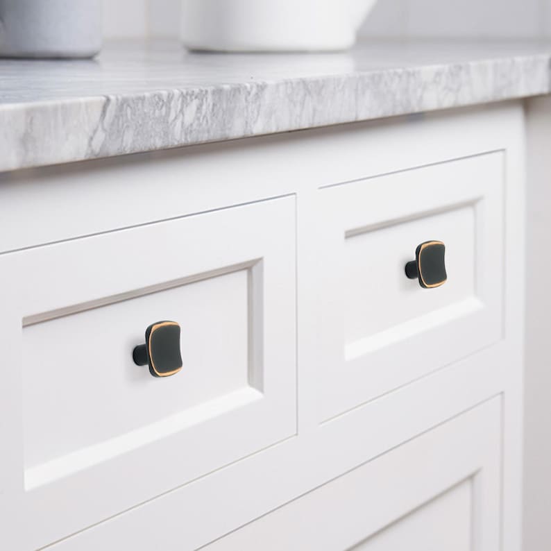 May include: Close-up of white cabinet doors with square black and gold cabinet knobs. The knobs have a dark, matte finish with a gold trim. The cabinet doors are set against a marble countertop.