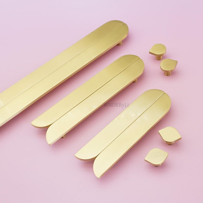 May include: A selection of gold-toned cabinet hardware, featuring long, curved handles and smaller knobs. The handles have a split design and come in different sizes. The hardware is displayed on a pale pink surface, creating a contemporary look.