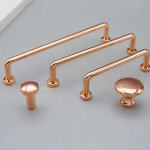 May include: A set of rose gold cabinet handles and knobs. The set includes two bar handles and two knob handles. The bar handles have a rounded, tubular design. The knobs are a mushroom style. The hardware is displayed on a grey surface.