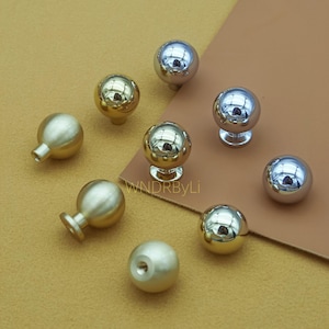 Peut inclure: Un assortiment de huit boutons de meubles sphériques en finitions or et argent. Certains boutons ont une tige pour le montage, tandis que d'autres sont des sphères pleines. Les boutons sont disposés sur une surface marron. Le texte "WNDRByLi" est visible.