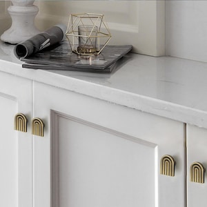 Op de afbeelding: Witte kastdeuren met goudkleurige, gewelfde ladeknopen. Een gouden geometrische kandelaar en een opgerold tijdschrift liggen op het aanrecht boven de kasten. De kastdeuren zijn effen en hebben een eenvoudig ontwerp.