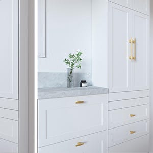 Op de afbeelding: Witte kasten met goudkleurige handgrepen. Het ontwerp is modern, met een marmeren aanrecht. Een heldere vaas met groen en een kleine zwarte pot staan op het aanrecht. De kastdeuren hebben verticale gouden handgrepen.