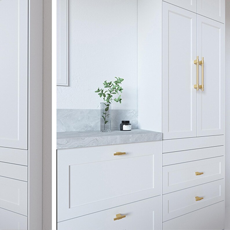 Op de afbeelding: Witte kasten met goudkleurige handgrepen. Het ontwerp is modern, met een marmeren aanrecht. Een heldere vaas met groen en een kleine zwarte pot staan op het aanrecht. De kastdeuren hebben verticale gouden handgrepen.
