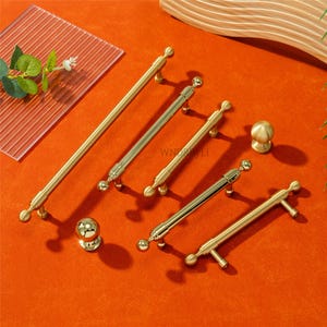 May include: A collection of gold-toned cabinet hardware, including various sizes of handles and knobs. The handles have a sleek, modern design with rounded ends and decorative details. The hardware is arranged on an orange surface.