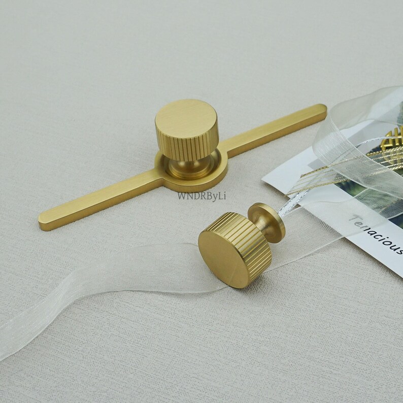 May include: Two gold-coloured cabinet knobs with a cylindrical design and ridged detailing. One knob is attached to a flat bar, and the other is separate. The knobs are on a light-coloured surface with a tag that says "Tenacious".
