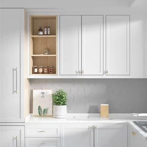 Op de afbeelding: Een moderne witte keuken met boven- en onderkasten, voorzien van gouden hardware. Een kleine plant in een pot en een ingelijst kunstwerk staan op het aanrecht. Open planken tonen potten en flessen.