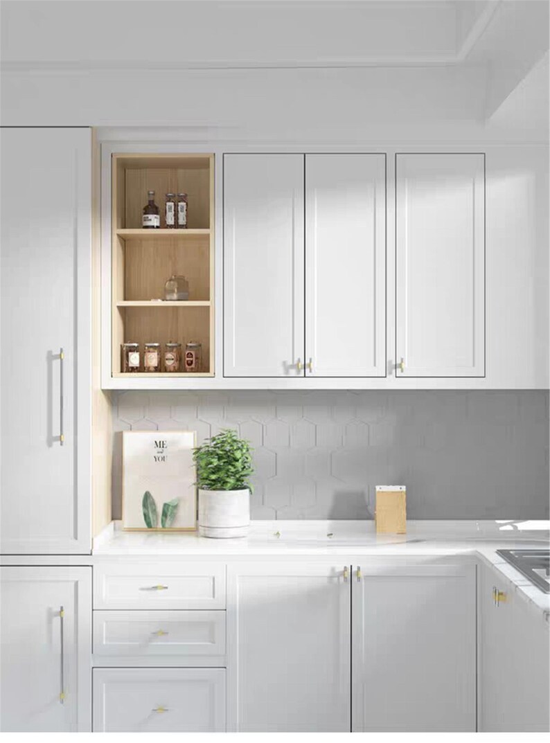 Op de afbeelding: Een moderne witte keuken met boven- en onderkasten, voorzien van gouden hardware. Een kleine plant in een pot en een ingelijst kunstwerk staan op het aanrecht. Open planken tonen potten en flessen.