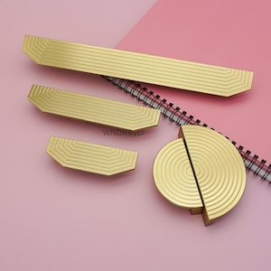 Op de afbeelding: Een verzameling goudkleurige kastbeslagen, waaronder rechthoekige en halfronde handgrepen. De handgrepen hebben een gegroefd, geometrisch ontwerp. De hardware wordt gepresenteerd op een roze ondergrond met een spiraalgebonden notitieboek.