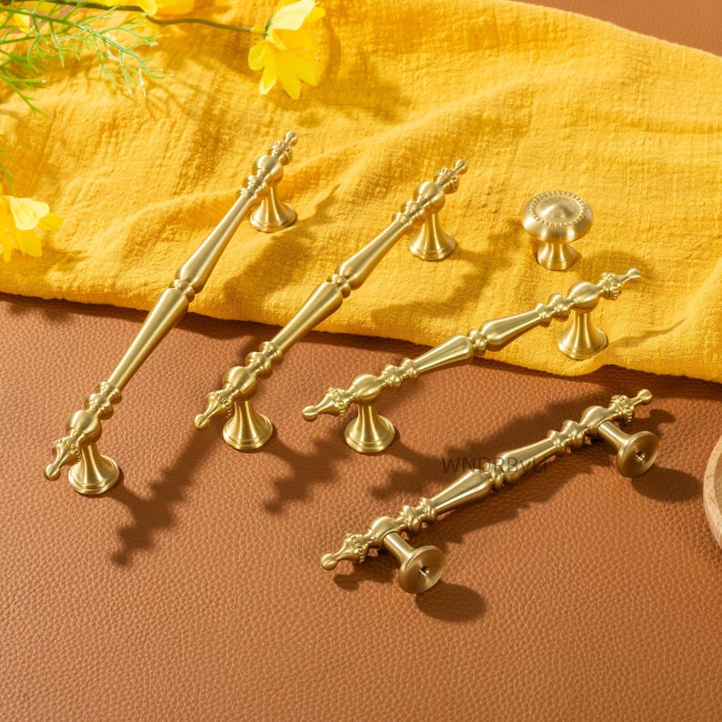 Peut inclure: Ensemble de quincaillerie de meubles en laiton de style antique, comprenant des poign&eacute;es et un bouton. Les poign&eacute;es pr&eacute;sentent des d&eacute;tails orn&eacute;s et sont pr&eacute;sent&eacute;es sur un tissu jaune. Id&eacute;al pour la d&eacute;coration int&eacute;rieure.