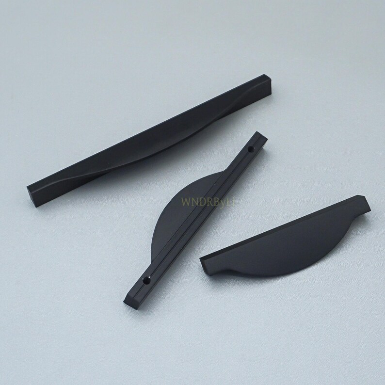 May include: Three matte black cabinet pulls in different shapes. One is a long, straight bar, another is a curved handle with screw holes, and the third is a smaller, curved pull. The pulls are on a light gray surface.