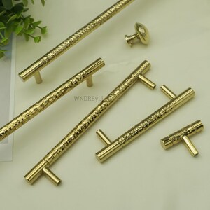 May include: A collection of gold-colored cabinet hardware, including handles and a knob. The handles have a textured, hammered appearance, and the knob has a rounded, oval shape. The hardware is designed for use on cabinets and drawers.