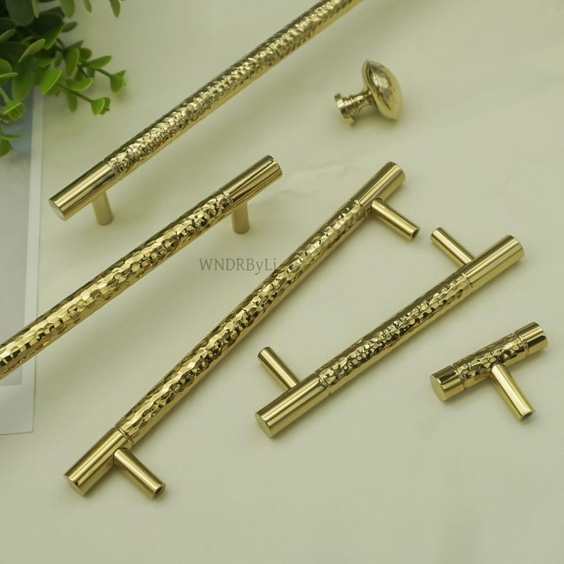 May include: A collection of gold-colored cabinet hardware, including handles and a knob. The handles have a textured, hammered appearance, and the knob has a rounded, oval shape. The hardware is designed for use on cabinets and drawers.