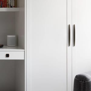May include: White cabinet doors with dark bronze-coloured, vertical bar handles. A small, square knob of the same colour is on a white drawer. A grey speaker sits on a shelf next to the cabinet.