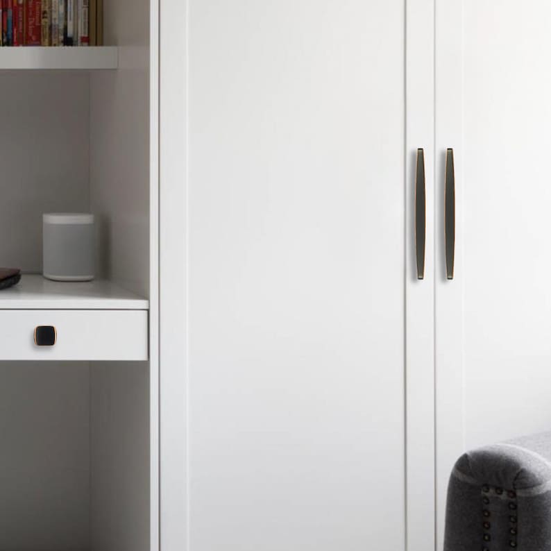 May include: White cabinet doors with dark bronze-coloured, vertical bar handles. A small, square knob of the same colour is on a white drawer. A grey speaker sits on a shelf next to the cabinet.