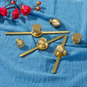 May include: A selection of gold-coloured cabinet hardware, including knobs and pulls, displayed on blue fabric. The hardware features various designs, including cylindrical and spherical shapes, suitable for furniture or cabinetry.