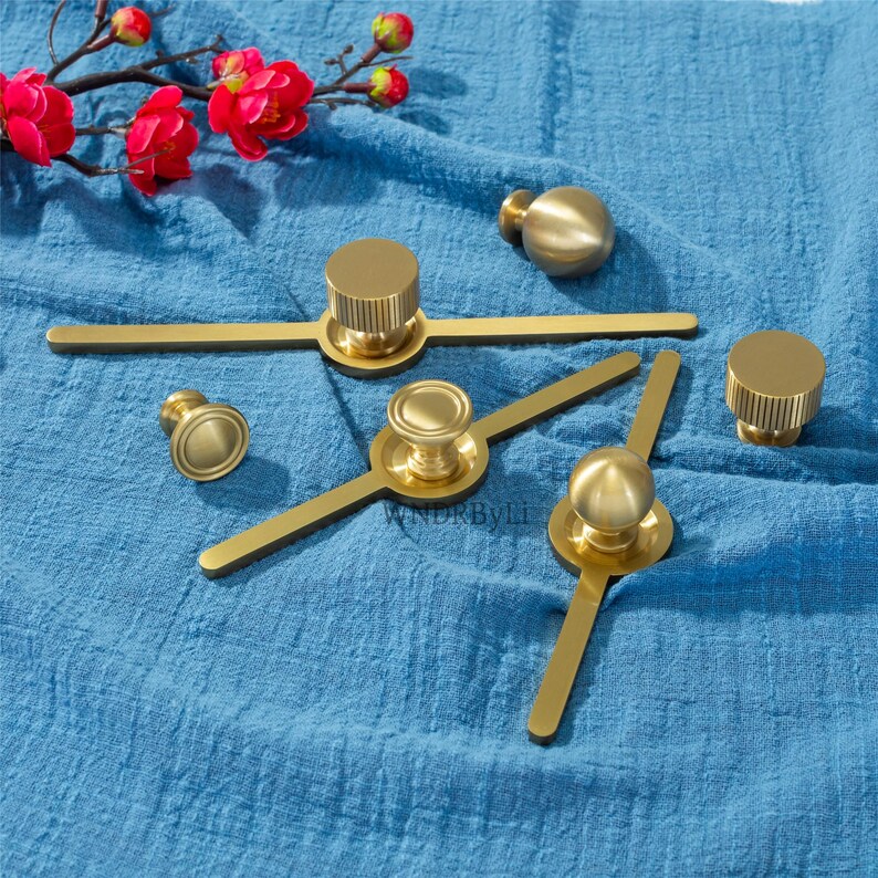 May include: A selection of gold-coloured cabinet hardware, including knobs and pulls, displayed on blue fabric. The hardware features various designs, including cylindrical and spherical shapes, suitable for furniture or cabinetry.