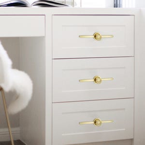 May include: A white cabinet with three drawers, each fitted with a gold-coloured handle. The handles feature a circular centre and a straight bar extending outwards. The cabinet is part of a larger white desk, with a chair visible.