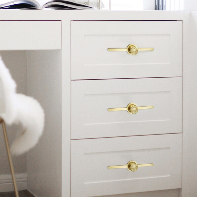 May include: A white cabinet with three drawers, each fitted with a gold-coloured handle. The handles feature a circular centre and a straight bar extending outwards. The cabinet is part of a larger white desk, with a chair visible.