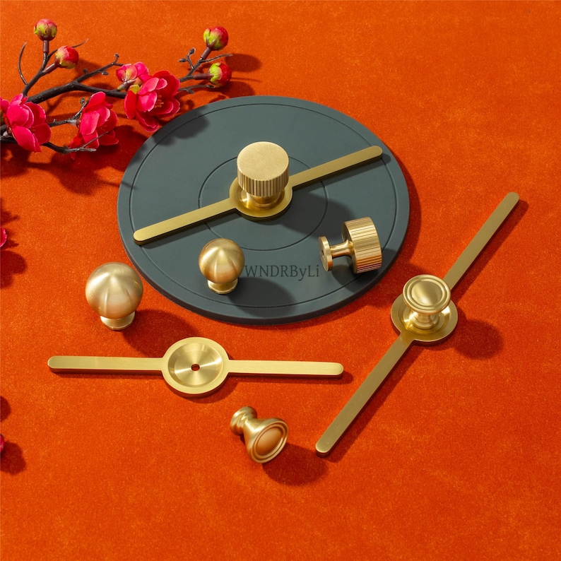 May include: A collection of brass cabinet hardware, including knobs, pulls, and a circular accent piece. The hardware is displayed on an orange surface with a dark gray circular accent piece. Red flowers are in the background.