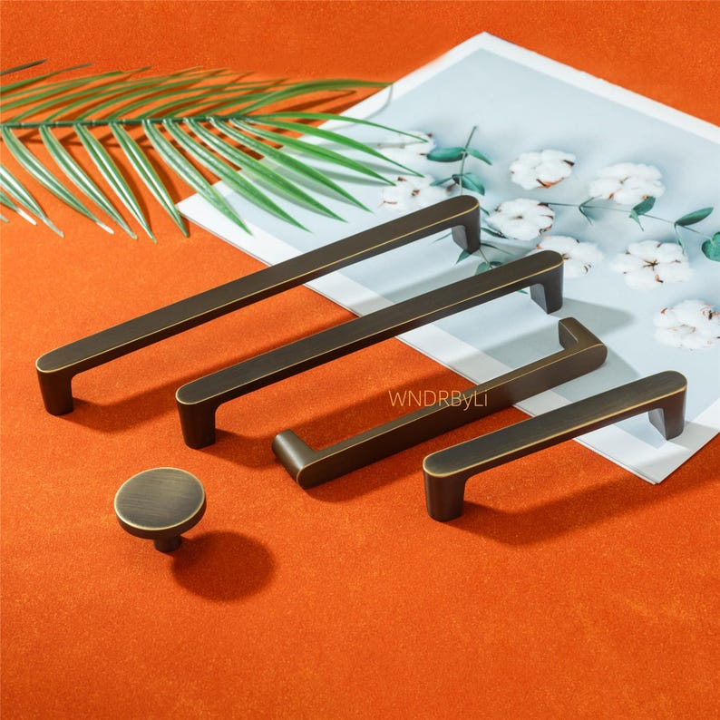 May include: A collection of bronze-toned cabinet hardware, including several rectangular bar pulls and a round knob. The hardware is arranged on an orange surface with a decorative leaf and floral background. The text "WNDRByLI" is visible.