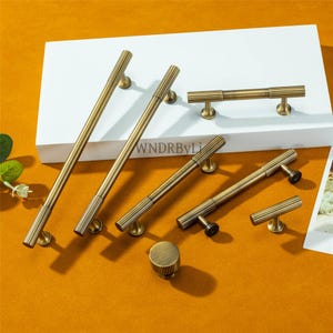 Puede incluir: Un conjunto de herrajes de lat&oacute;n antiguo para armarios, que incluye manijas de varios tama&ntilde;os y un pomo. Las manijas tienen un dise&ntilde;o acanalado y se exhiben sobre una superficie blanca contra un fondo naranja c&aacute;lido. El texto "WNDRByLi" es visible.