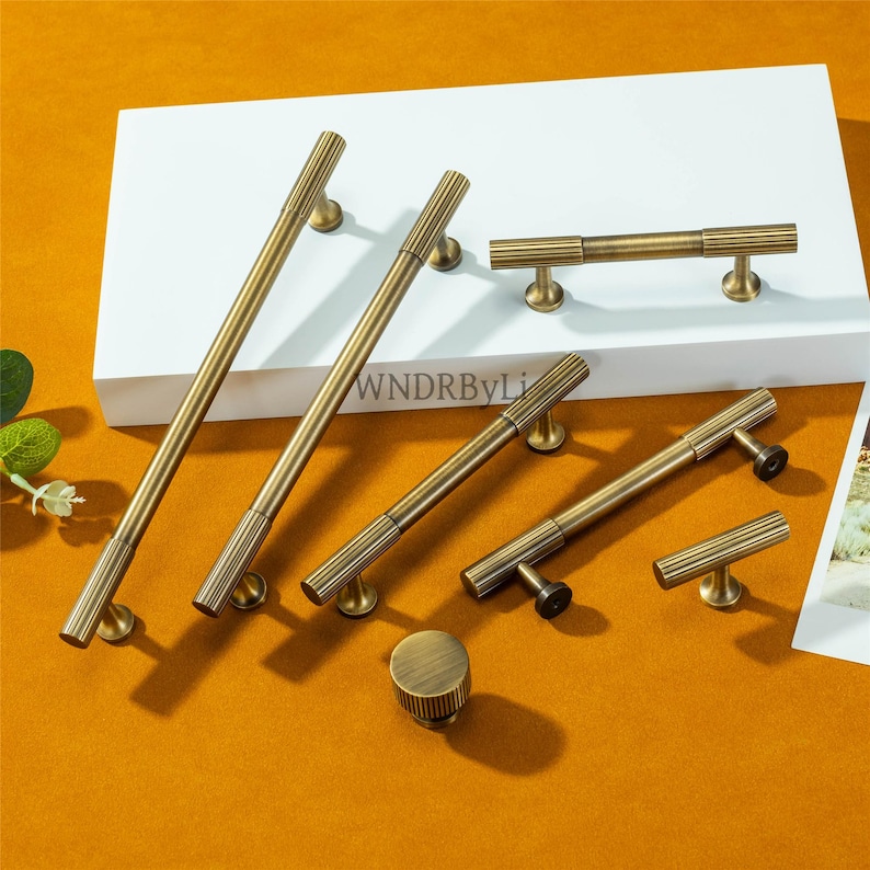 Puede incluir: Un conjunto de herrajes de lat&oacute;n antiguo para armarios, que incluye manijas de varios tama&ntilde;os y un pomo. Las manijas tienen un dise&ntilde;o acanalado y se exhiben sobre una superficie blanca contra un fondo naranja c&aacute;lido. El texto "WNDRByLi" es visible.